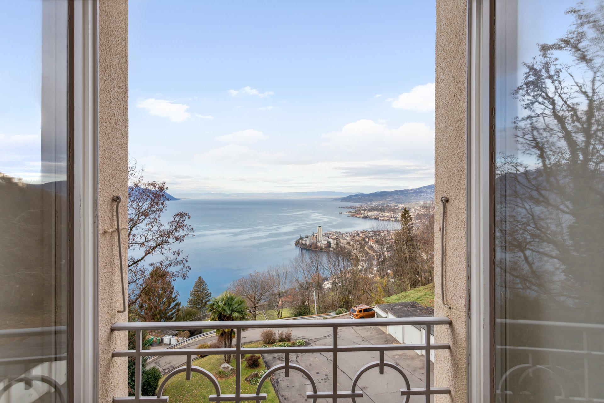 La vue d’un lac depuis un balcon, avec des eaux calmes, des nuages légers et des arbres longeant le rivage au loin, sur un fond de ciel bleu doux.