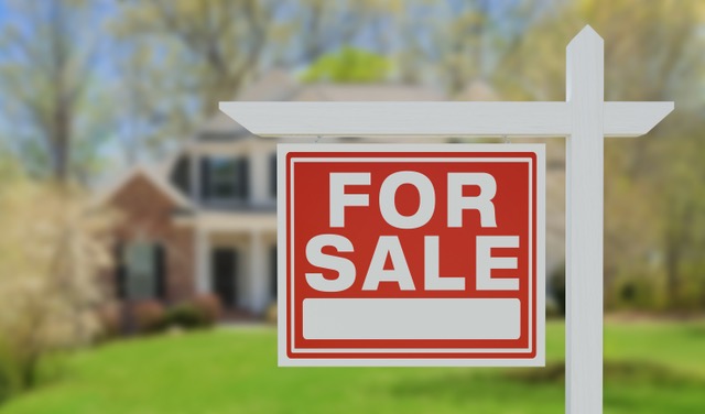 Au premier plan, un piquet blanc porte un panneau rouge « À VENDRE ». En arrière-plan, une maison apparaît floue